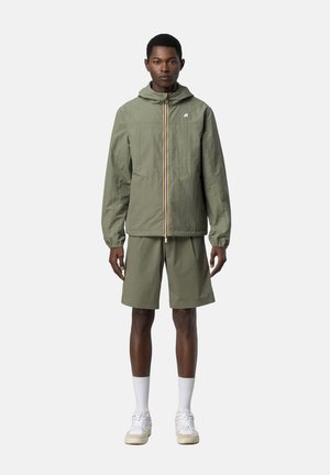 Giovane uomo in piedi che indossa giacca con cappuccio verde oliva e pantaloncini abbinati con calzini bianchi e scarpe da ginnastica su sfondo semplice.