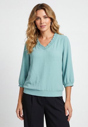 Vrouw met golvend blond haar, die een lichtblauwe blouse met lange mouwen en een gerimpelde V-hals en een zwarte broek draagt, staand tegen een effen achtergrond.