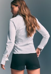 Chemise blanche à manches longues avec une coupe ajustée et détail aux poignets, associée à un short noir. Tissu lisse, design minimaliste, sans motifs apparents.