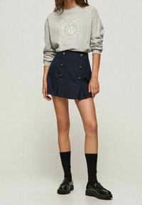 Šedá mikina s logem, v kombinaci s námořnickou sukní se zlatými knoflíky, sklady a strukturovaným střihem. Černé ponožky a lesklé boty.