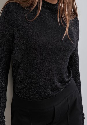 Zbliżenie osoby noszącej czarną, błyszczącą bluzkę z długim rękawem i czarne spodnie, z widocznymi jasnobrązowymi włosami.