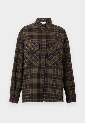 Høj person iført solbriller, ternet skjorte over pink top, marineblå bukser med knapper i siden, holder en stor mørk tekstureret tote taske.