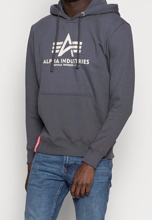 Mężczyzna noszący szarą bluzę z kapturem Alpha Industries z logo i napisem, w zestawieniu z niebieskimi dżinsami, stoi na tle jednolitego jasnego tła.
