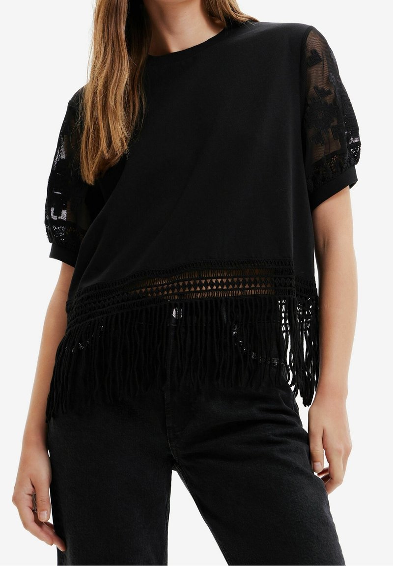 Černý top s krátkými, průhlednými texturovanými rukávy, vyznačující se háčkovaným lemem a dlouhým třásňovým detailem na spodní části.