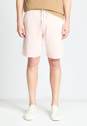 Mann trägt hellrosa knielange Shorts, beiges T-Shirt und weiße Schnürsneaker, steht vor einem schlichten weißen Hintergrund.
