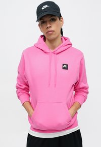 Roza kapucna majica s sprednim žepom, vrvicama in črnim kvadratnim logotipom Nike Air. Obuta s črno kapo in plastno belo majico.