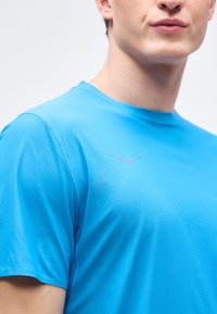 Κοντινό πλάνο ενός ατόμου που φοράει ένα ζωηρό μπλε υφασμάτινο κοντομάνικο πουκάμισο με ένα μικρό, μωβ λογότυπο πουλιού στο στήθος.