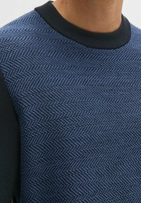 Blaues Sweatshirt mit strukturiertem Fischgrätenmuster, schwarzen einfarbigen Ärmeln und einem schwarzen gerippten Kragen. Fokus auf Details des gewebten Stoffes.