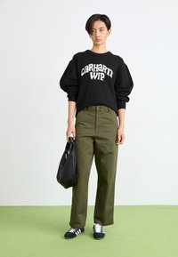 Melnas pārslēgtas dūrainas svārkas ar baltu "Carhartt WIP" logotipu, olīvu zaļi plaša kāju bikses, melna soma un zili sporta apavi ar baltām svītrām.
