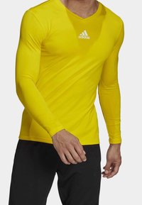 Man i en åtsittande långärmad gul Adidas-tröja och svarta byxor, sedd från halsen till mitt lår mot en vit bakgrund.