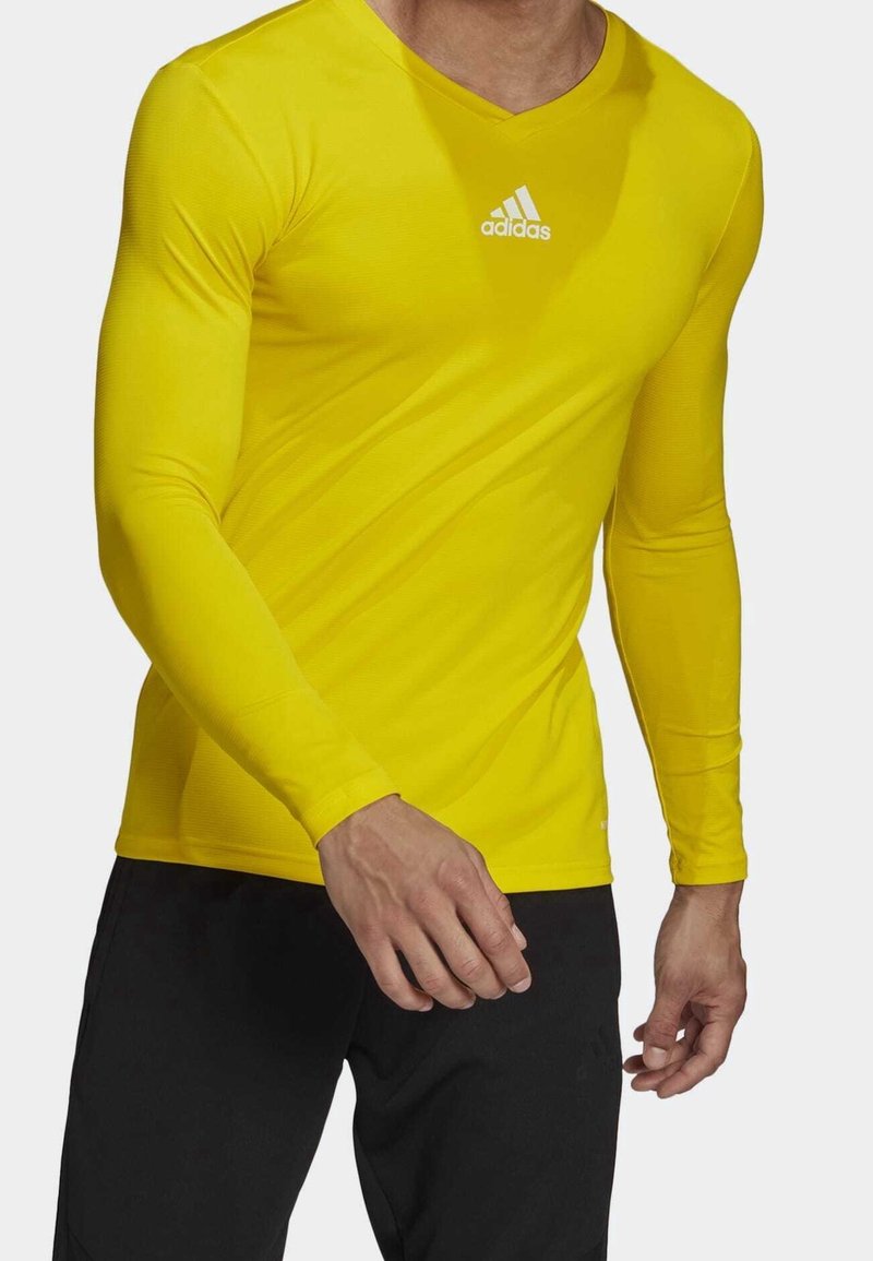 Man i en åtsittande långärmad gul Adidas-tröja och svarta byxor, sedd från halsen till mitt lår mot en vit bakgrund.