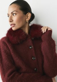 Bordeaux wollen mix cardigan met een pluizige nepbont kraag, twee voorzakken en grote zwarte knopen. Lange, geribde mouwen met een gebreide textuur.