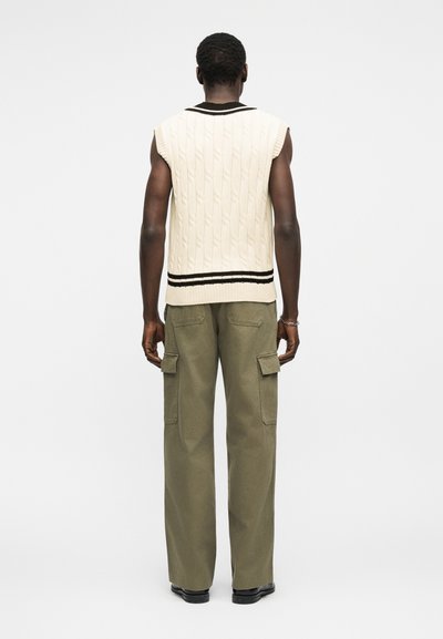 Άντρας που φαίνεται από πίσω φοράει αμάνικο κρεμ πλεκτό πουλόβερ με μαύρες ρίγες και χακί παντελόνι cargo με μαύρα παπούτσια.