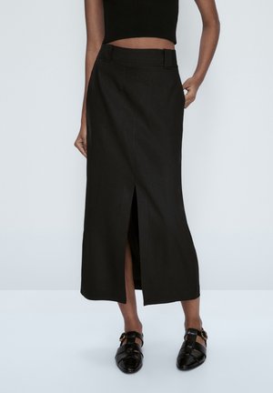 Femme portant une jupe midi noire taille haute avec une fente à l'avant, des chaussures noires à boucle, et un haut court noir sans manches, debout sur un fond blanc.