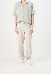 Světle zelená košile s krátkým rukávem a knoflíkovým zapínáním, v kombinaci s béžovými lněnými kalhotami s elastickým pasem a bílými teniskami.