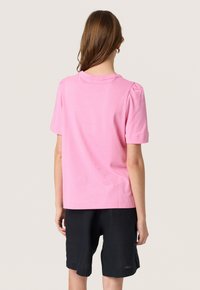 Lyserød bomulds T-shirt med korte puffede ærmer og rund hals, parret med mørke linnedshorts, set fra bagsiden.