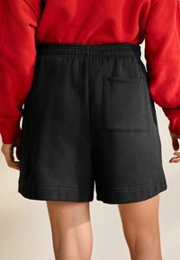 Sorte shorts med elastisk taljebånd, der har en enkelt laplomme på bagsiden og en glat tekstur. Sættes sammen med en rød top.