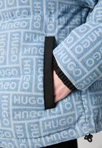 Jasnoniebieska jeansowa kurtka z wzorem logo HUGO, z czarną lamówką kieszeni i elastycznymi ściągaczami. Teksturowana tkanina o casualowym designie.