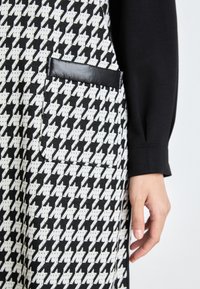 Mustavalkoinen houndstooth-kuosisella mekolla, jossa on kiiltävä musta taskuyksityiskohta ja pitkähihaisella yläosalla, näkyy käsi lepäämässä kankaalla.