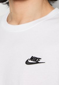 White cotton t-shirt featuring a black embroidered Nike logo on the left chest area. Crew neck design with set-in sleeves.