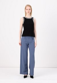 Top senza maniche nero con bordi bianchi, abbinato a pantaloni blu a gamba larga e scarpe nere con punta affilata, creando un outfit a contrasto.
