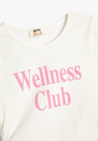 Hvid ribbet bomulds T-shirt med teksten "Wellness Club" i fed pink skrift. Rund hals og korte ærmer.