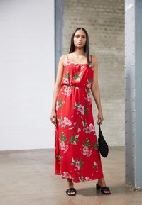 Vestido maxi floral vermelho com alças finas, corte solto, cintura franzida, apresentando flores cor-de-rosa e folhas verdes. Combinado com uma bolsa preta.