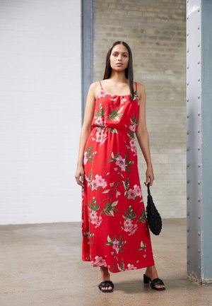 Vestido maxi floral vermelho com alças finas, corte solto, cintura franzida, apresentando flores cor-de-rosa e folhas verdes. Combinado com uma bolsa preta.