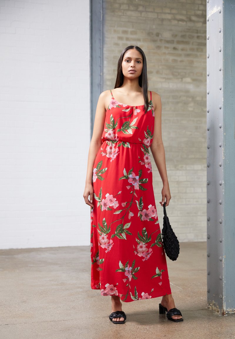 Vestido maxi floral vermelho com alças finas, corte solto, cintura franzida, apresentando flores cor-de-rosa e folhas verdes. Combinado com uma bolsa preta.