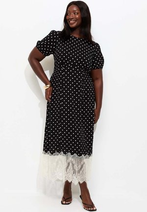 Kobieta w czarnej sukience midi w białe kropki z białą koronkową lamówką, stojąca z ręką na biodrze i uśmiechająca się, na stopach czarne sandały.