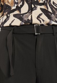 Pantalons noirs taille haute avec une texture lisse et une ceinture avec une boucle rectangulaire en argent, assortis à un chemisier à motifs.