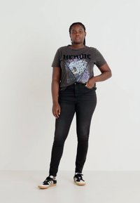 Črna grafična majica s kratkimi rokavi z napisom "HEROIC" in leopardskim vzorcem; v kombinaciji s črnimi oprijetimi jeans hlačami in črnimi supergami z belimi detajli.