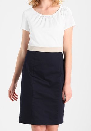 Frau trägt eine weiße Kurzarmbluse mit beigem Taillenbund und einem knielangen dunkelblauen Bleistiftrock, steht vor weißem Hintergrund.