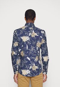 Camicia blu navy con motivi floreali in beige chiaro e bianco, maniche lunghe e colletto classico. Presenta un orlo curvo e dettagli con bottoni.