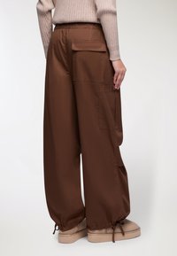 Pantaloni a gamba larga marroni realizzati in tessuto liscio, con tasche laterali e polsini elasticizzati. La vita è di tipo arricciato per maggiore comfort.
