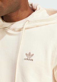 Cream-colored hoodie made of soft fabric, featuring an embroidered brown Adidas logo on the chest and a drawstring hood.