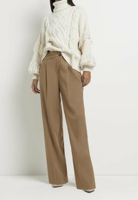 Pull crème en maille torsadée avec manches à franges, associé à un pantalon marron taille haute à jambes larges avec plis sur le devant et bouton latéral.