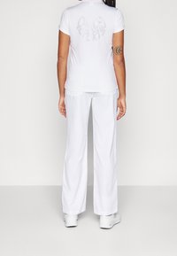 Bílá polo košile s krátkým rukávem a reflexním motivem na zádech, v kombinaci s volnými bílými kalhotami a bílými sportovními botami.