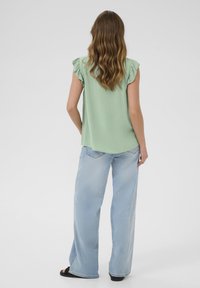 Un haut vert menthe avec des manches à volants, associé à un jean bleu clair à jambes larges et des sandales noires, présenté de dos contre un fond uni.