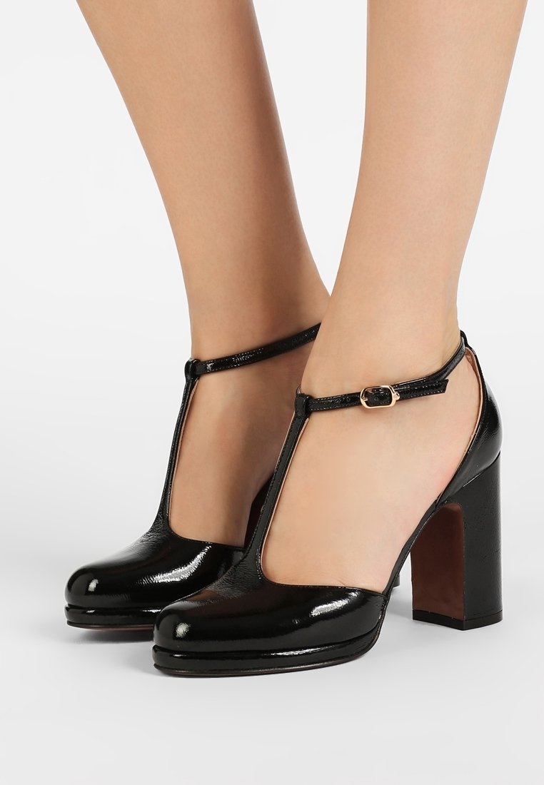 Chaussures à talons hauts en cuir verni noir avec bride en T et boucle autour de la cheville, portées pieds nus sur fond blanc.
