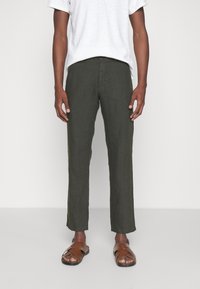 Pantaloni in lino verde scuro con un design a gamba dritta, abbinati a una maglietta grigia chiara e sandali marroni, indossati da una figura maschile in piedi.