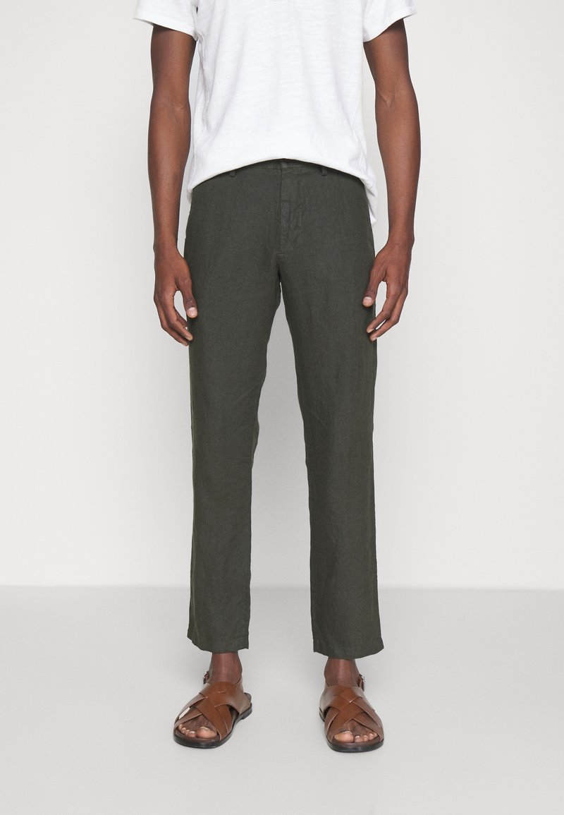 Pantaloni in lino verde scuro con un design a gamba dritta, abbinati a una maglietta grigia chiara e sandali marroni, indossati da una figura maschile in piedi.