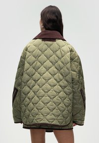 Person med langt mørkt hår iført en overdimensioneret kviltet olivengrøn jakke med brun krave og lynlåsedetaljer, der vender væk mod en ensfarvet baggrund.
