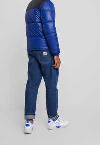 Blå puffjacka med svarta axlar, denimjeans och vita sneakers med blå accenter. Jackan har en kviltad textur och elastiska ärmslut.