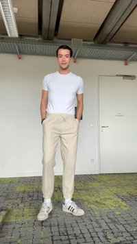 Jeune homme aux cheveux foncés debout à l'intérieur, portant un t-shirt blanc, un pantalon beige et des baskets blanches sur un sol gris et vert à motifs.