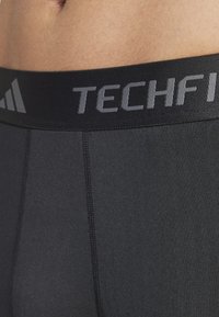 Pantaloni sportivi neri con finitura opaca, dotati di una ampia vita elastica con la scritta "TECHFIT" in grigio e una vestibilità aderente.