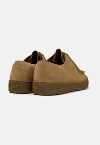 Zapatos de gamuza marrón con forma redondeada, suela de goma triturada texturizada y cordones mínimos, con un logotipo grabado en el talón.