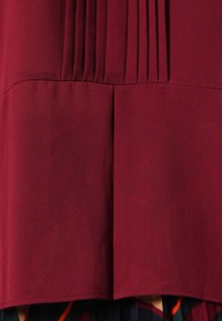 Chemisier bordeaux avec des plis verticaux, texture lisse et ourlet droit. Le tissu semble léger et fluide.