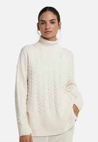 Kobieta ubrana w sweter z dekoltem typu stójka w kolorze off-white, w plecionkę, z długimi rękawami, stoi z jedną ręką w kieszeni na tle jednolitego białego tła.