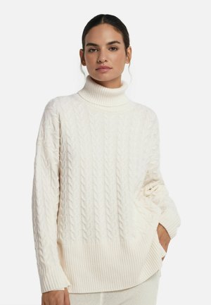 Kvinna iförd en off-white kabelstickad polo med långa ärmar, stående med en hand i fickan mot en plain vit bakgrund.